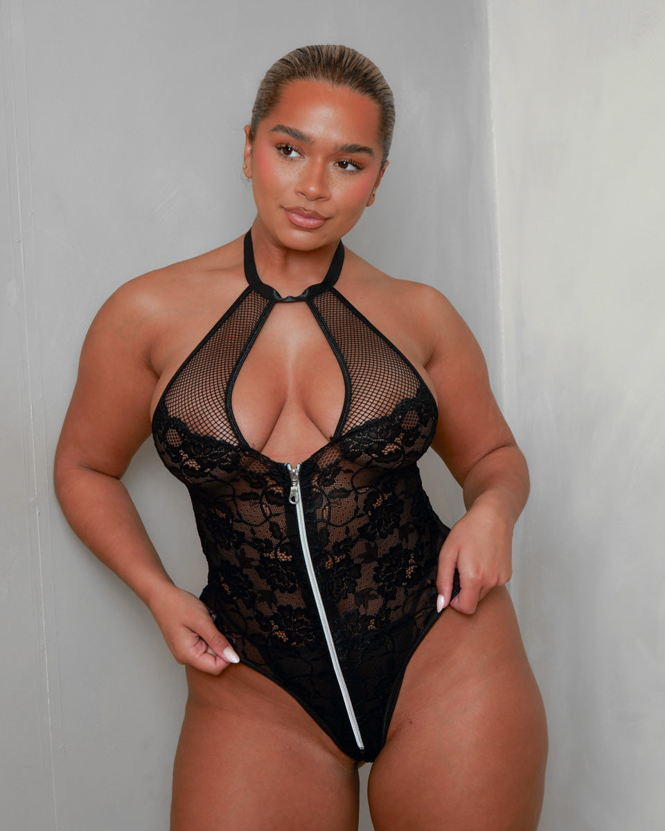 Front view of the Tutti Boudoir Zuri Bodysuit in Onyx Black, combining a plunging halterneck, sheer fishnet panels, floral lace and statement zip fastening for bold, confidence-boosting lingerie.