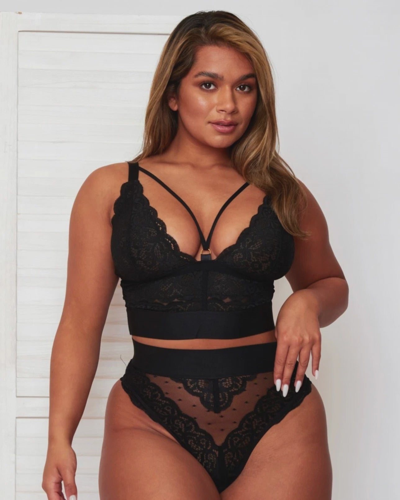Model turning to show Tutti Rouge’s black lace bralette and matching high-waisted briefs, featuring a strappy plunge neckline, wide underband and sheer lace detailing.